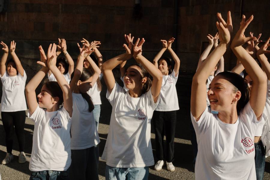 Eine Gruppe von Mädchen und Frauen in weißen T-Shirts und dunklen Hosen, die gerade tanzen und dabei die Arme in die Luft heben.