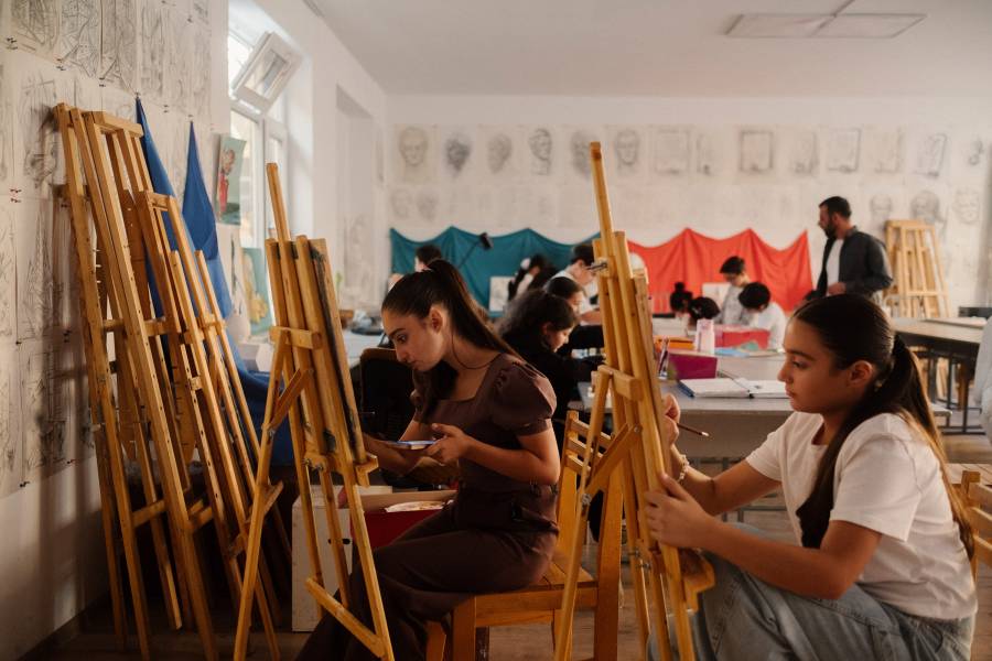 Zwei Mädchen, die im Malclub vor einer hölzernen Staffelei sitzen und gerade mit Pinsel und Farben arbeiten.