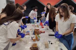 Eine Gruppe von jungen Frauen und Männer beim Vorbereiten der Mahlzeiten für Armeeangehörige. Sie stehen um einen großen Tisch mit weißer Tischdecke und tragen alle lila Handschuhe für diese Arbeit.