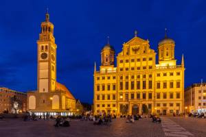 Das Bild zeigt das Augsburger Rathaus und den Perlachturm am Abend. Die beiden Gebäude leuchten in ockerfarbenen und warmgelben Tönen vor einem dunkelblauen Abendhimmel, davor sitzen viele Menschen auf dem Boden.