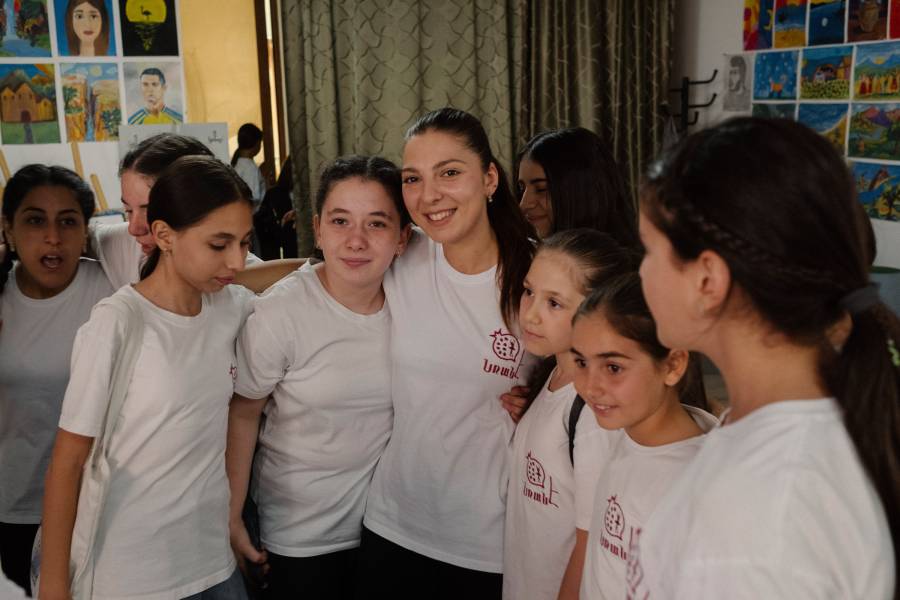 Die Trainer Anush steht in der Mitte, rechts und links von ihr ein Teil der Teilnehmerinnen des Tanzclubs. Sie tragen alle weiße T-Shirts, haben die dunklen Haare zusammengebunden und legen sich die Arme um die Schultern.