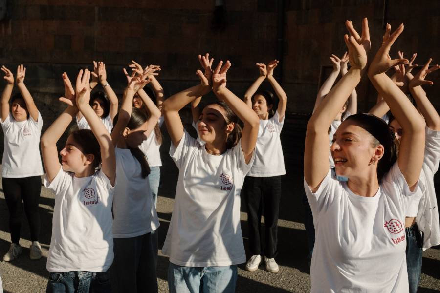 Anush, die Tanztrainerin und ihre Mädchen tanzen mit erhobenen Händen und alle mit dem Kopf nach rechts gewandt im Sonnenlicht.