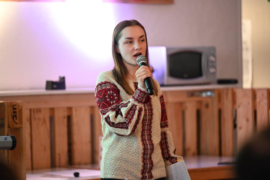 Anna Sobolevska aus der Ukraine während ihres Vortrags, mit einem Mikrofon in der Hand. Sie hat lange hellbrauen Haare, trägt eine Brille und eine traditionelle Bluse aus ihrer Heimat.