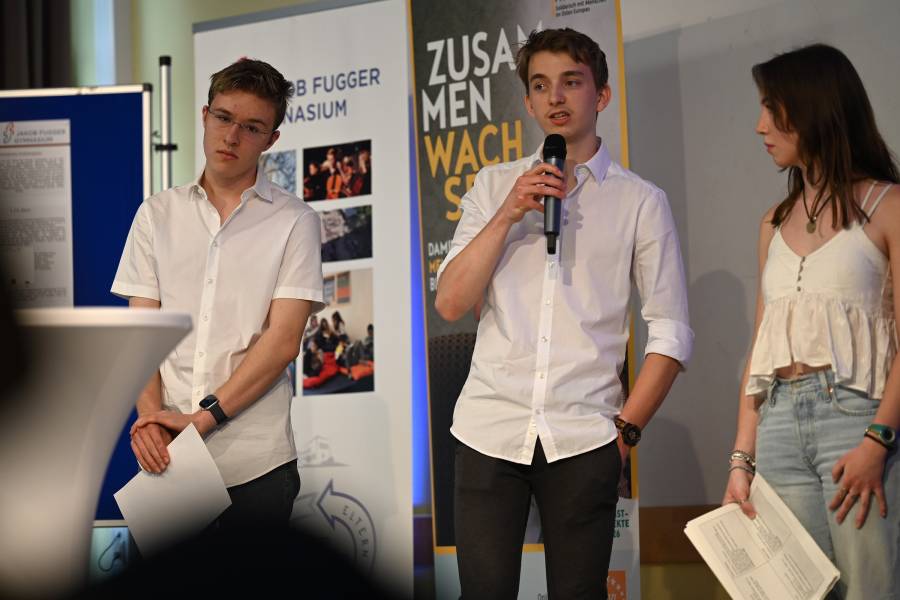 Zwei junge Schüler stehen im weißen Hemd auf dem Podium ihrer Schule und stellen Fragen, daneben steht eine junge Frau im hellen T-Shirt und mit einem Zettel in der Hand.