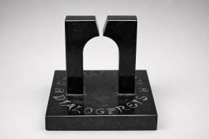 Zu sehen ist eine schwarze, glänzende Skulptur auf einem quadratischen Sockel vor hellem, neutralem Hintergrund. Zwei hohe, rechteckige Säulen stehen parallel nebeneinander und sind im oberen inneren Bereich halbkreisförmig ausgeschnitten. Zusammen bilden die Aussparungen eine große, bogenartige Öffnung in der Mitte. Auf dem Sockel ist in heller Schrift das Wort „DIALOGPREIS“ eingraviert.