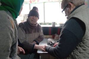 In einem hellen Raum sitzt ein älterer Mann mit Mütze und Pullover an einem Tisch. Eine Pflegerin mit Brille hält seinen Arm; auf dem Tisch liegen medizinische Utensilien. Links ist eine ältere Frau mit Kopftuch teilweise zu sehen.