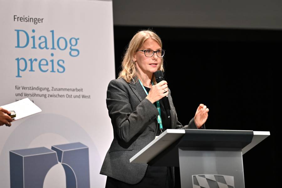 Aleksandra Królak-Wąsowicz steht an einem Rednerpult auf einer Bühne und spricht in ein Handmikrofon. Neben ihr ist ein Banner des Freisinger Dialogpreises zu sehen. Während ihres Vortrags gestikuliert sie mit einer Hand und stellt die Arbeit der Stiftung Kreisau für Europäische Verständigung vor.