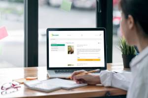 Ein Laptop auf einem hellen Holztisch zeigt den neuen, übersichtlichen Renovabis-Onlineshop. Auf dem Monitor ist das aktuelle Pfingstplakat 2026 zu sehen, daneben ein markanter gelber Button „In den Warenkorb legen“. Eine Person im Vordergrund deutet die einfache Bedienung am Arbeitsplatz an. Die Bildstimmung ist hell, aufgeräumt und modern.